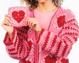 Una mujer muestra un cardigan tejido a mano, rosa y rojo, con un corazón de ganchillo. Este es un ejemplo de qué es un cardigan.