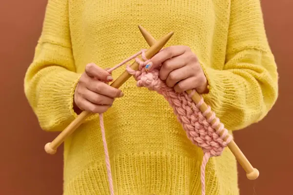 Manos tejiendo un suéter amarillo con hilo rosa, mostrando cómo hacer ochos de 4 puntos.