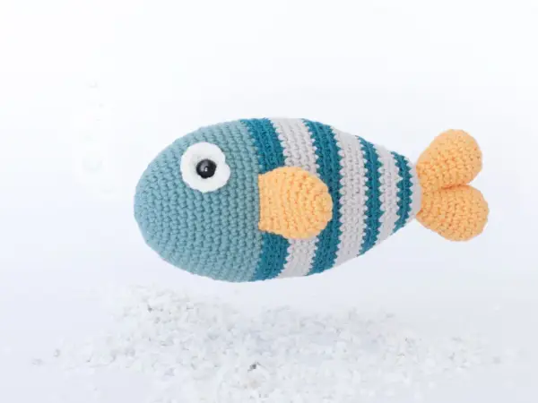 Un pez amigurumi a rayas azules y grises con aletas naranjas, flotando sobre guijarros blancos. ¡Encuentra aquí peces amigurumi patrón gratis!
