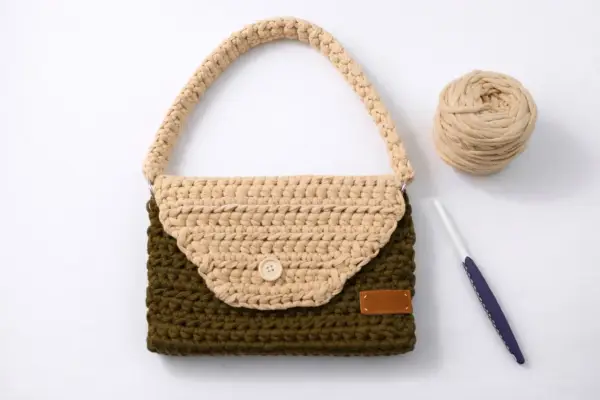 Bolso de trapillo bicolor, verde y beige, con asa y botón de madera. A su lado, ovillo de lana y ganchillo.