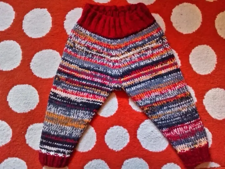 Pantalón de punto para bebé con rayas multicolores y cintura roja, sobre fondo de lunares.