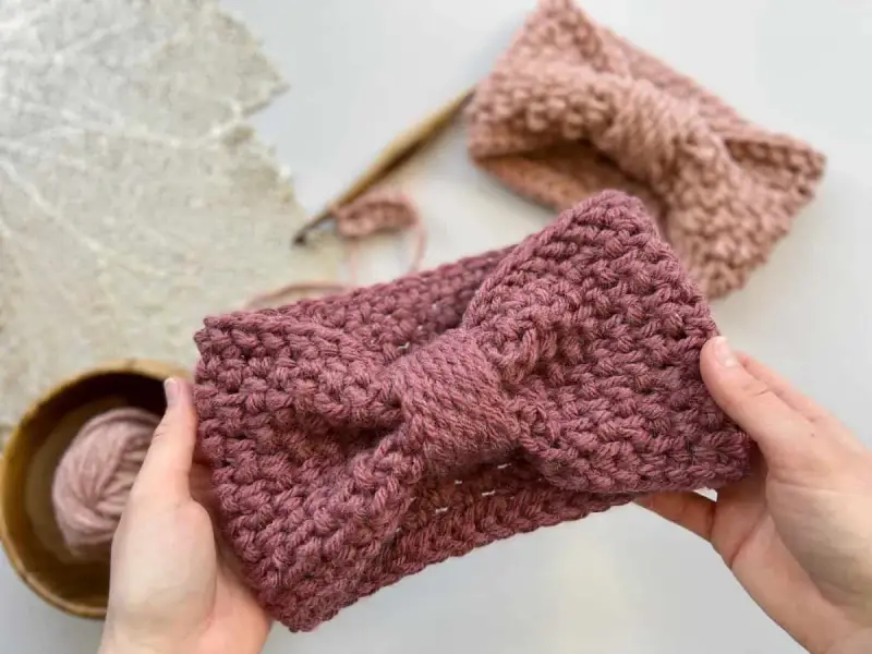 Manos sostienen una diadema tejida a crochet con un lazo. Detrás, otra diadema y ovillo de lana. ¡Descubre patrones gratis de diademas de ganchillo!