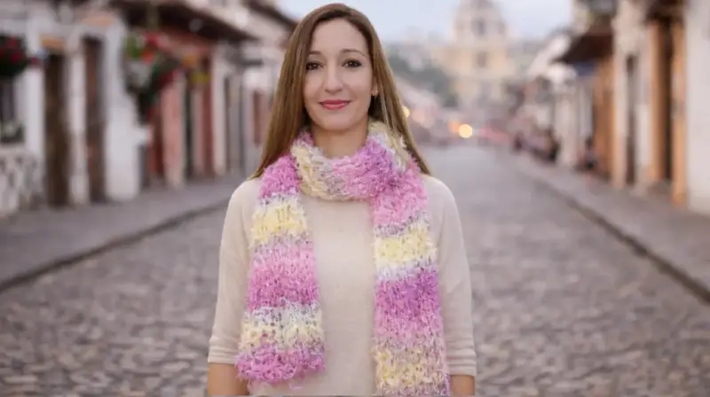 Mujer sonriente luce bufandas de dos agujas modernas en tonos pastel, paseando por una calle empedrada.